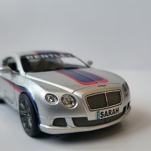 May include: A silver toy car model of a Bentley Continental GT with red and blue racing stripes and a number plate that reads "SARAH".