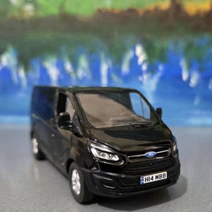May include: A black Ford Transit van model with silver wheels. The van has a detailed front grille, headlights, and a license plate that reads "HI4 MBB". The Ford logo is visible on the front.