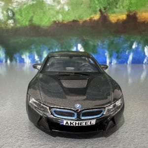 May include: A black BMW i8 model car with blue accents on the front. The car has a license plate that reads "AKHEEL". The car is on a gray surface with a colorful background.