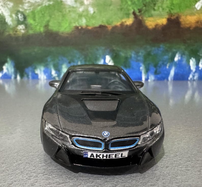 May include: A black BMW i8 model car with blue accents on the front. The car has a license plate that reads "AKHEEL". The car is on a gray surface with a colorful background.