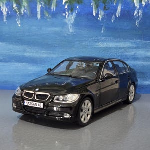 May include: A black sedan model with silver alloy wheels and a number plate reading "HASSAN 40". The car is on a grey surface with a blue and white painted backdrop.