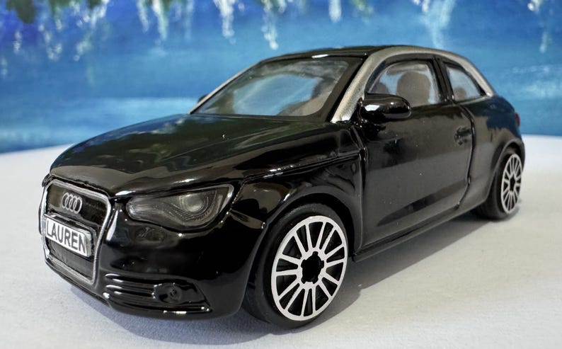 Peut inclure: Une voiture jouet noire avec des accents argent&eacute;s, avec une plaque d'immatriculation "LAUREN". La voiture a des roues d&eacute;taill&eacute;es et un design &eacute;pur&eacute;, ressemblant &agrave; une petite voiture de sport. Le fond est bleu flou.