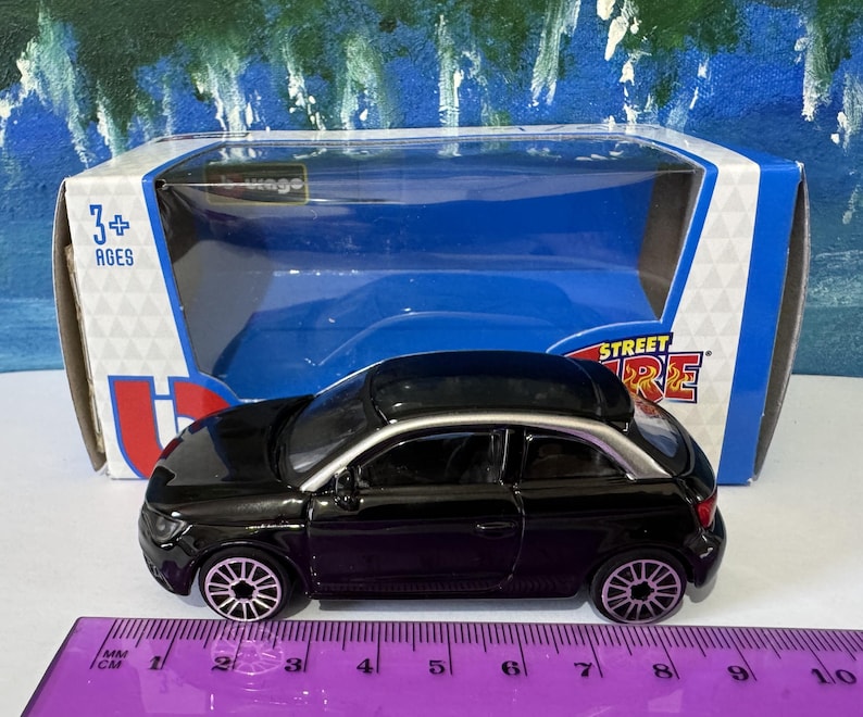 Peut inclure: Une voiture miniature noire avec des garnitures argent&eacute;es, pr&eacute;sent&eacute;e dans son emballage d'origine. La voiture jouet a des roues d&eacute;taill&eacute;es et un design &eacute;l&eacute;gant. L'emballage comprend le texte "STREET FIRE" et "3+ AGES". Une r&egrave;gle est au premier plan.