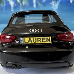 Peut inclure: Une voiture mod&egrave;le Audi A1 noire avec des feux arri&egrave;re rouges. La plaque d'immatriculation affiche "LAUREN" en jaune. Le logo Audi est visible au-dessus.