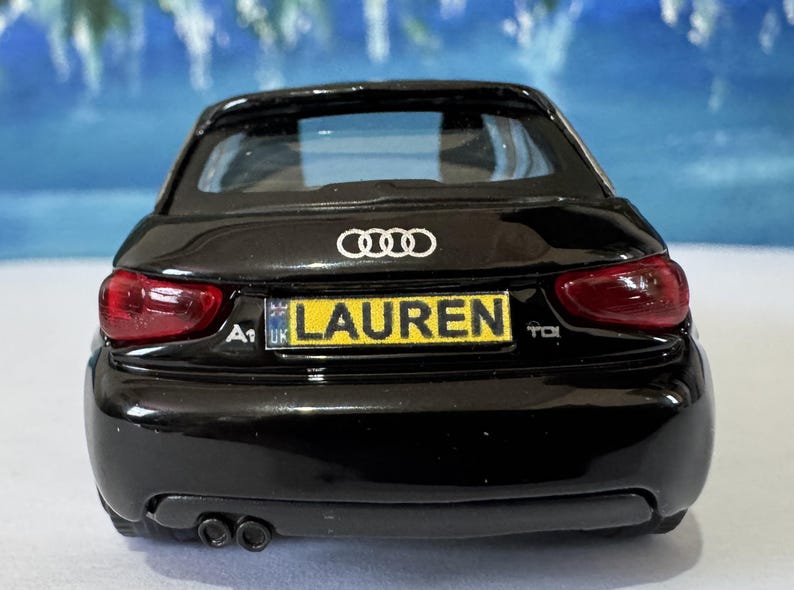 Peut inclure: Une voiture mod&egrave;le Audi A1 noire avec des feux arri&egrave;re rouges. La plaque d'immatriculation affiche "LAUREN" en jaune. Le logo Audi est visible au-dessus.