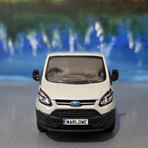 Puede incluir: Maqueta de furgoneta Ford Transit blanca con parachoques delantero y espejos laterales negros. El modelo tiene la palabra "MARLOWE" en la matrícula y un logotipo azul de Ford en la parte delantera.