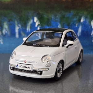 Pode incluir: Um carro em miniatura Fiat 500 branco com teto preto, rodas prateadas e uma placa dianteira com o texto JORDAN. O veículo possui faróis redondos, um acabamento cromado no para-choque dianteiro e um pequeno emblema Fiat no capô.
