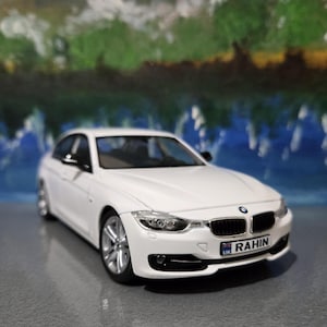 May include: A white model car with a black roof and silver rims. The car has a license plate that reads "RAHIN" and the UK flag. The car is set against a blurred background of blue and green.