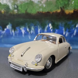 May include: A cream-coloured vintage Porsche model car with black tyres and chrome accents. The model has a curved windscreen and the word "PEMBROKE" is on the front number plate. The background is a blurred painting of a landscape.