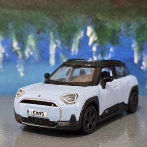May include: A pale blue toy car with a black roof and wheels. The car has a number plate that reads "LEWIS". This is a small-scale model, possibly a collectible or a child's toy.