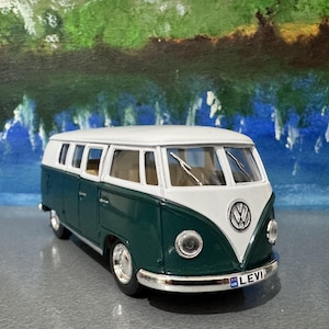 May include: A detailed model of a green and white Volkswagen Type 2 bus. The model has a white roof, green body, and chrome detailing. The front features the VW logo and a license plate that reads "LEVI UK".