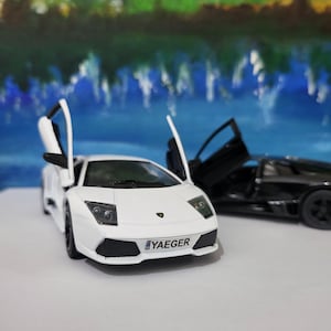 Könnte beinhalten: Ein weißes Lamborghini Spielzeugauto mit geöffneten Türen und einem schwarzen Nummernschild mit der Aufschrift "YAEGER". Das Auto steht auf einer blauen und grünen Oberfläche.