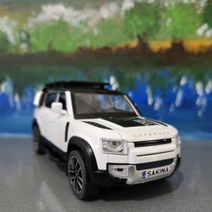 May include: A white and black toy SUV with the word "DEFENDER" printed on the hood. The toy car has black tires and a roof rack. The license plate reads "SAKINA". The background is a blurred image of a landscape.