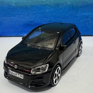 May include: A black toy car with the name "MARTIN" on the number plate. The car has a shiny finish, white wheels, and a red detail on the front grille. The background is a blue gradient.