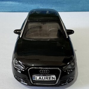 Peut inclure: Une voiture jouet noire avec le nom "LAUREN" sur la plaque d'immatriculation. La voiture a une finition brillante, des d&eacute;tails argent&eacute;s et le logo Audi &agrave; l'avant. C'est une maquette d'une voiture moderne.