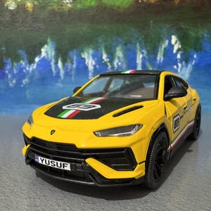 Könnte beinhalten: Ein gelbes Spielzeugauto mit schwarzen Akzenten und der Nummer "63" an der Seite. Die Motorhaube hat einen schwarzen Streifen mit grünen, weißen und roten Streifen. Das Nummernschild lautet "YUSUF".