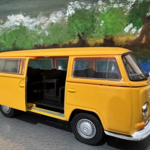 May include: A yellow toy van with an open side door, revealing black interior seating. The van has a white bumper and silver wheels. The background features a painted landscape with trees and water.