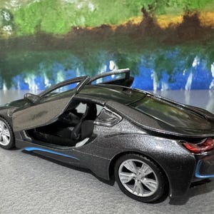 May include: A dark gray toy car with the driver's side door open. The car has silver wheels and blue accents. The background features a colorful abstract painting.