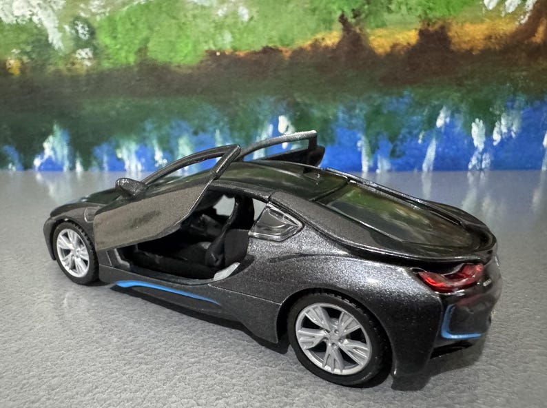 May include: A dark gray toy car with the driver's side door open. The car has silver wheels and blue accents. The background features a colorful abstract painting.