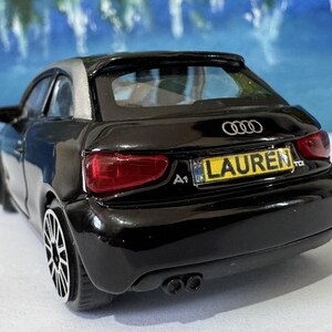 Peut inclure: Une maquette de voiture Audi A1 noire avec un toit argent&eacute;. La voiture a une plaque d'immatriculation qui indique "LAUREN". La voiture a des feux arri&egrave;re rouges et des roues noires. Le logo Audi est visible &agrave; l'arri&egrave;re.