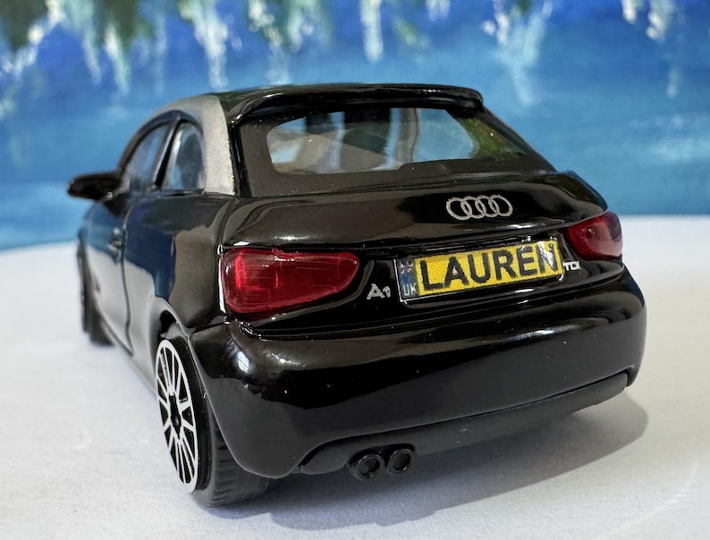 Peut inclure: Une maquette de voiture Audi A1 noire avec un toit argent&eacute;. La voiture a une plaque d'immatriculation qui indique "LAUREN". La voiture a des feux arri&egrave;re rouges et des roues noires. Le logo Audi est visible &agrave; l'arri&egrave;re.