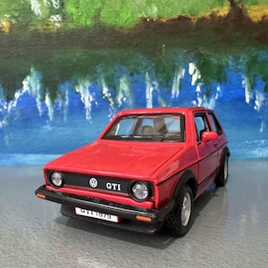 以下が含まれることがあります： 前面に「GTI」の文字が入った赤いモデルカー。車は黒いトリム、シルバーのホイール、そして「GTI 1979」と書かれたナンバープレートが付いています。背景は青と緑で描かれたシーンです。