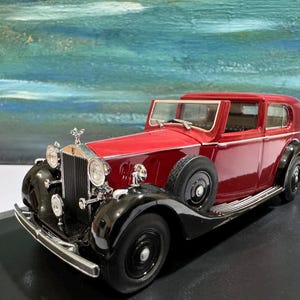 May include: A red vintage Rolls Royce Phantom II model car with black accents and chrome details. The car is on a black display base.