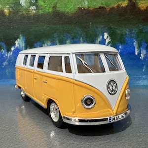 Peut inclure: Un modèle réduit de combi Volkswagen jaune et blanc avec des détails chromés. Le véhicule a un toit blanc, une carrosserie jaune et le logo Volkswagen. La plaque d'immatriculation indique "MALIK".