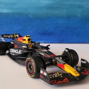 Puede incluir: Modelo detallado de un coche de carreras de Fórmula 1. El coche es principalmente negro con detalles en rojo y amarillo, con logotipos de patrocinadores como "Oracle" y "Red Bull". El alerón delantero muestra "SYBT Red" y "Claro".