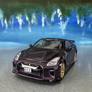May include: A miniature dark purple sports car with gold-colored wheels. The car has a spoiler and a license plate that reads "KYLE". The background features a blurred blue and green landscape.