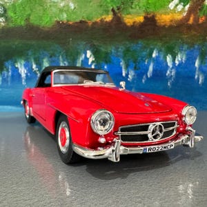 May include: A red classic convertible car model with a black roof. The car has chrome detailing, a detailed front grille, and a number plate. The background features a painted landscape with blue and green hues.