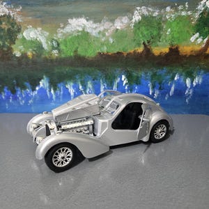 Può includere: Un'auto modello argentata con il cofano aperto, che rivela il motore. L'auto ha un design classico con parafanghi arrotondati e una carrozzeria aerodinamica. Le ruote hanno dettagli a raggi intricati. Lo sfondo presenta un paesaggio dipinto.