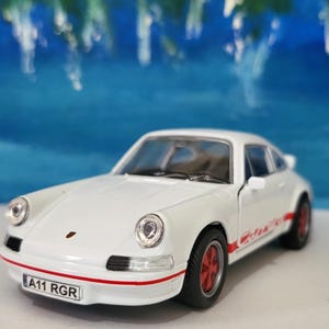 Peut inclure: Modèle de voiture jouet blanc d'une Porsche 911 avec des bandes de course rouges et une plaque d'immatriculation qui indique "A11 RGR".