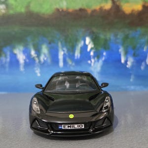Puede incluir: Un coche deportivo de juguete negro con la matrícula "EMILIO". El coche tiene un diseño elegante con faros y un pequeño emblema verde. El fondo es una imagen borrosa de un paisaje azul y verde.