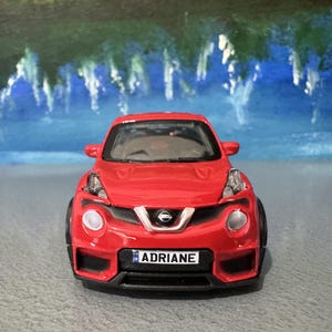 Op de afbeelding: Een rode speelgoedauto met zwarte accenten, met een Nissan-embleem en de naam "ADRIANE" op de nummerplaat. De auto heeft ronde koplampen en een sportief ontwerp, tegen een vage achtergrond.