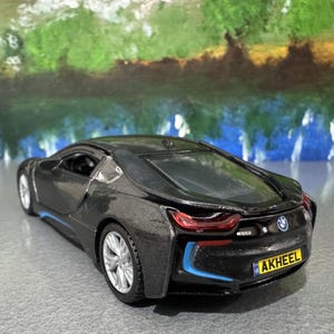 May include: A black BMW i8 toy car with silver rims and blue accents. The toy car has a license plate that reads "AKHEEL". The background is a blurred painting of green and blue colors.