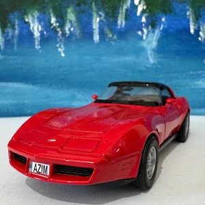 Puede incluir: Un coche modelo Corvette rojo de los años 70 con techo negro y una matrícula que dice "AZIM".