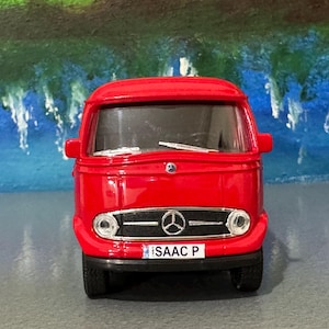 May include: A vibrant red toy Mercedes-Benz van with a black grill and tyres. The van has a number plate that reads "ISAAC P". The background is a painted scene of blue water and green trees.