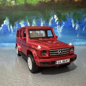 Op de afbeelding: Een rode speelgoed-SUV met een zwarte grille en het woord "GILBERT" op de nummerplaat. De speelgoedauto heeft een gedetailleerd ontwerp, inclusief koplampen, zijspiegels en een Mercedes-Benz logo.