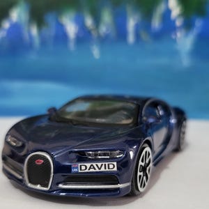 May include: A dark blue toy car with a sleek design, featuring a black front grill and silver accents. The car has the name "DAVID" on the license plate. The wheels are silver and black. The background is a blurred blue.