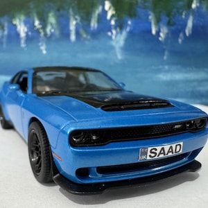 May include: A blue Dodge Challenger model car with a black hood and black wheels. The car has a black grill and black headlights. The car has a license plate that says "SAAD" with a UK flag on it.