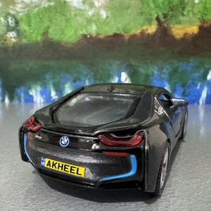 May include: A black BMW i8 model car with blue accent lines and red taillights. The toy car has a license plate that reads "AKHEEL". The BMW logo is visible on the rear of the car.