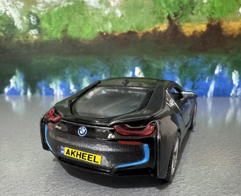 May include: A black BMW i8 model car with blue accent lines and red taillights. The toy car has a license plate that reads "AKHEEL". The BMW logo is visible on the rear of the car.