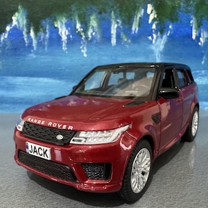 Puede incluir: Un modelo de coche Range Rover Sport rojo con techo negro y ruedas plateadas. La matrícula delantera dice "JACK". El coche está sobre un fondo pintado de azul y verde.