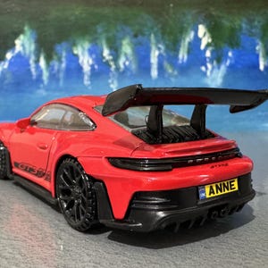 May include: A red Porsche GT3 RS model car with black wheels and a large rear wing. The car has a black rear bumper and a license plate that reads "ANNE". The car is set against a blue and white background.