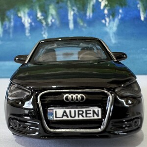 Peut inclure: Une voiture jouet noire avec une plaque d'immatriculation indiquant "LAUREN". La voiture a une finition brillante et une calandre avant d&eacute;taill&eacute;e avec le logo Audi. L'arri&egrave;re-plan est une image floue d'un paysage bleu et vert.