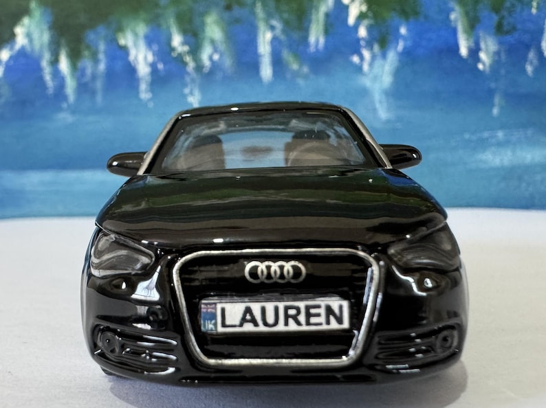 Peut inclure: Une voiture jouet noire avec une plaque d'immatriculation indiquant "LAUREN". La voiture a une finition brillante et une calandre avant d&eacute;taill&eacute;e avec le logo Audi. L'arri&egrave;re-plan est une image floue d'un paysage bleu et vert.