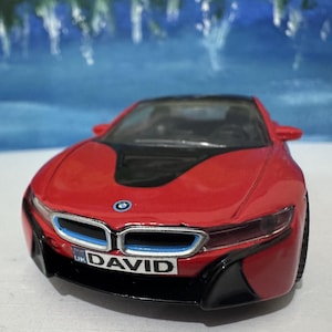 Op de afbeelding: Rode speelgoedauto met een zwart dak en een blauwe en zilveren grille. De auto heeft een kentekenplaat met de tekst "UK DAVID".