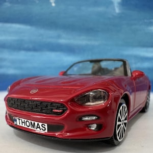May include: A red toy convertible car with black and silver wheels. The car has a black grill and the word "THOMAS" on the number plate. The car is set against a blue and white background.