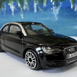 Peut inclure: Une voiture jouet Audi A1 noire avec un toit argent&eacute; et des roues blanches. La voiture porte le nom "LAUREN" sur sa plaque d'immatriculation. L'arri&egrave;re-plan est une image floue bleue et blanche.